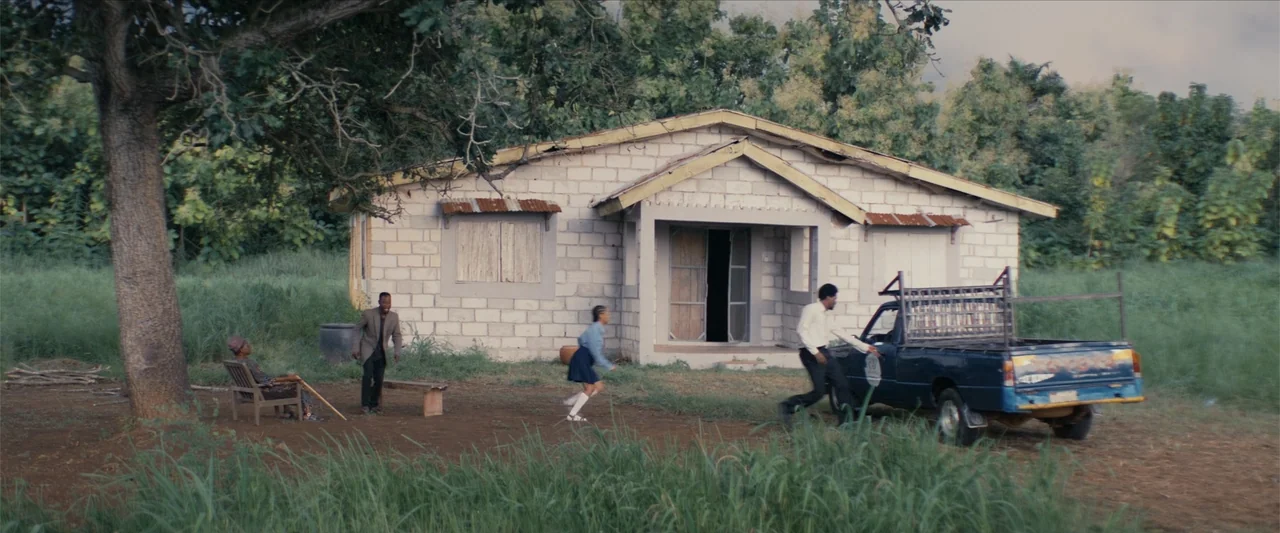 Ms. Kanyin (2025) movie still: wide — A modest cinder block home sits in a grassy clearing with a large tree. Four individuals are positioned around…