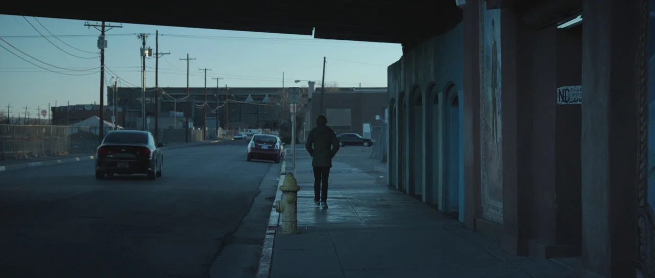 Frank (2014) movie still: wide — A quiet urban street stretches beneath a bridge underpass during the daytime. A lone figure walks along the…