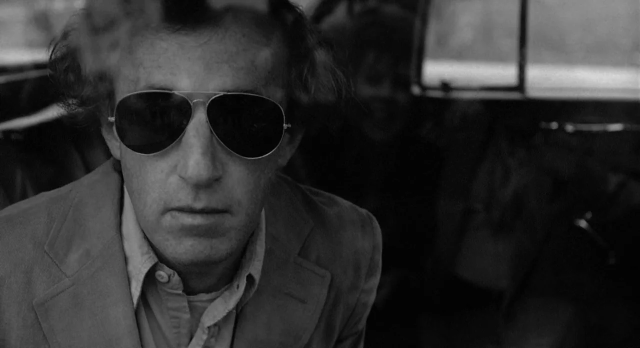 Stardust Memories (1980) movie still: closeup — Inside a car cabin with blurry window reflections visible in the background. A man in sunglasses…