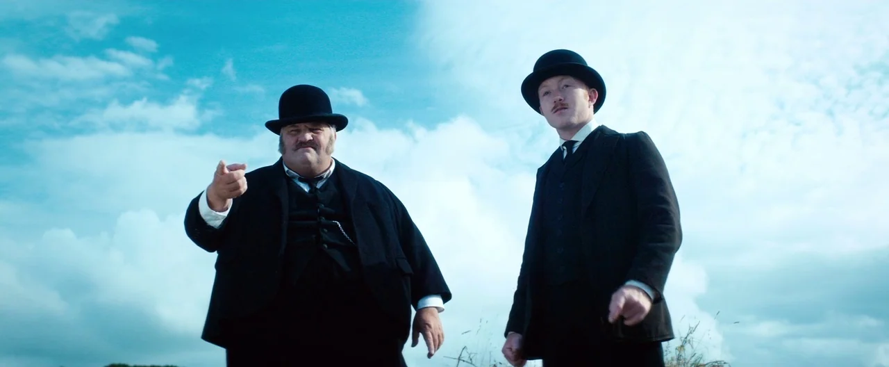 Slack Bay (2016) movie still: medium — Two men in vintage black suits stand against an expansive, bright blue, cloudy sky. Both figures look down at…