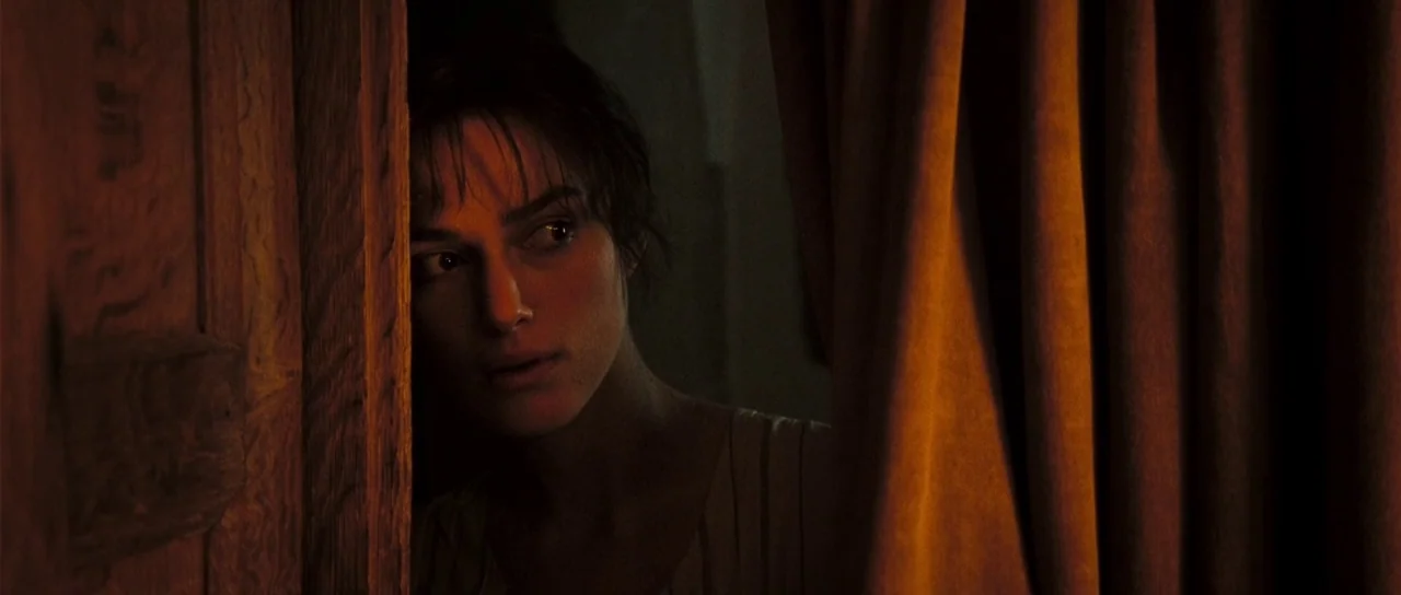 Pride & Prejudice (2005) movie still: closeup — A woman looks through a narrow opening between a thick curtain and a wooden frame. The low light…
