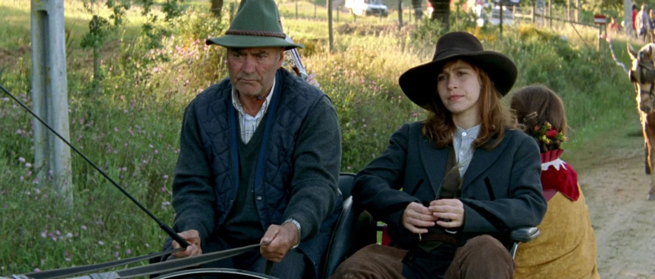 Arabian Nights: Volume 3 The Enchanted One (2015) movie still: medium — A man and woman ride in a carriage along a rural dirt road. The path is…