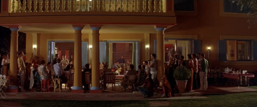 Talk To Her (2002) movie still: A wide, symmetrical shot capturing a bustling social gathering on the terrace of a Mediterranean villa at night. The…