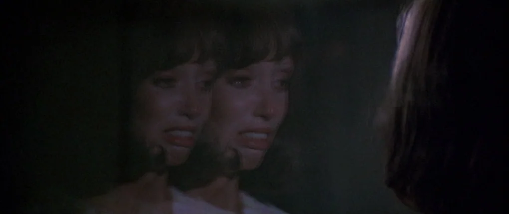 3 Women (1977) movie still: closeup — A dimly lit, shadowed bathroom features a wall-mounted mirror capturing a reflection. A woman with dark hair…