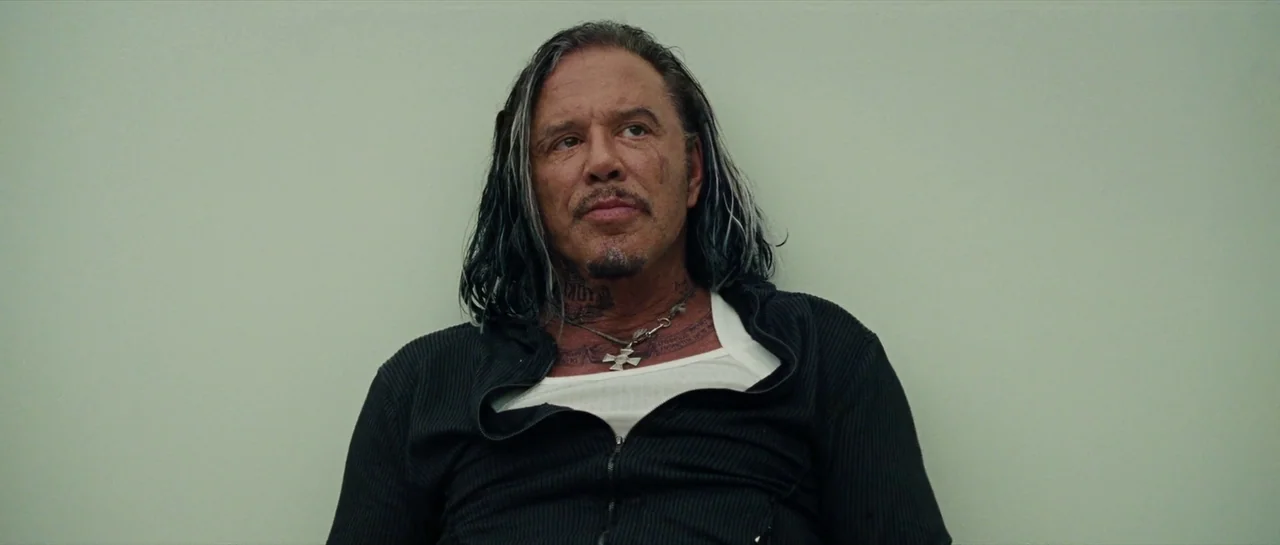 Iron Man 2 (2010) movie still: medium — A man sits against a plain, light green wall in an indoor space. The man has long hair and neck tattoos…