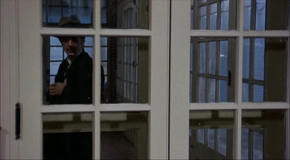 The King Of Marvin Gardens movie still: medium — A dark sunroom with large white-framed glass doors at night. A man in a suit stands behind the…