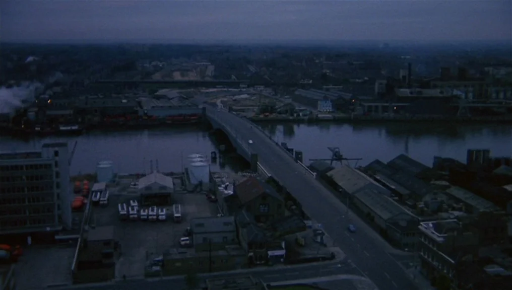 Performance (1970) movie still: establishing — An aerial view shows an industrial river district with warehouses, storage tanks, and a bridge. The…