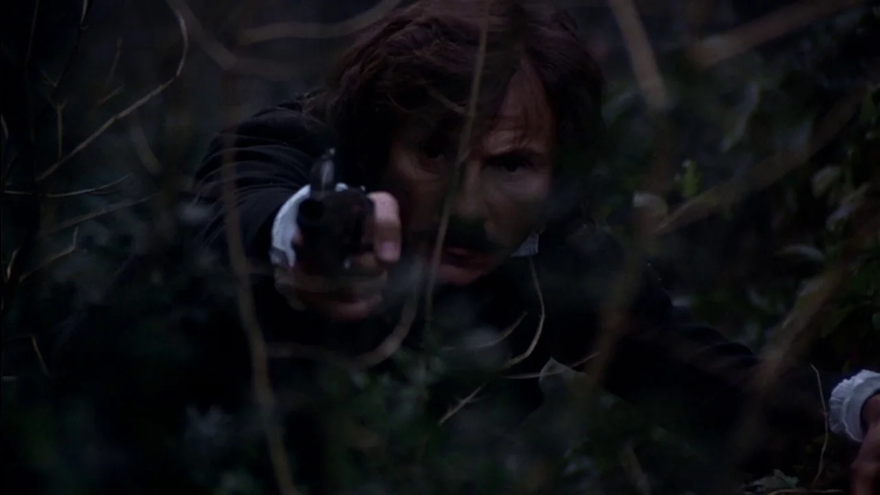 The Duellists (1977) movie still: medium — A man hides within dark, tangled brush at night. He aims a pistol directly forward while looking toward…