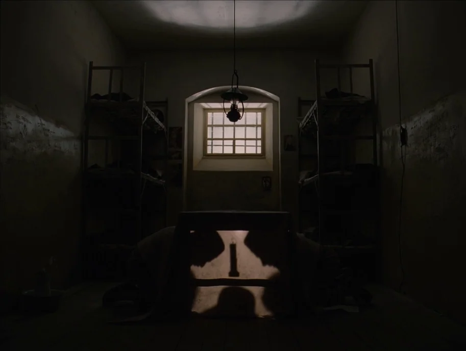 The Grand Budapest Hotel (2014) movie still: wide — A dark, spartan prison cell features two metal bunk beds against the side walls. A white sheet…