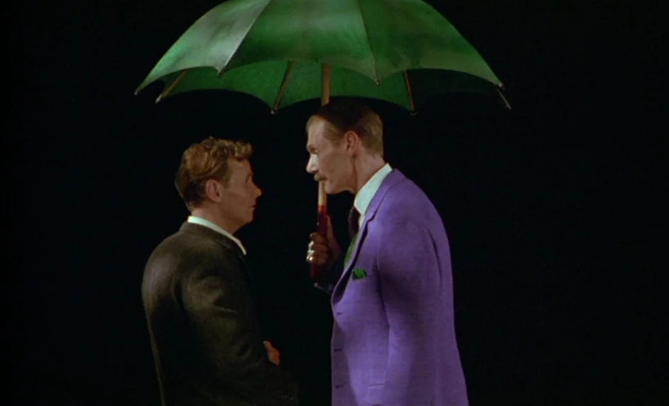 Wittgenstein (1993) movie still: medium — Two men stand together on a plain black stage under a green umbrella. The taller man wears a purple suit…