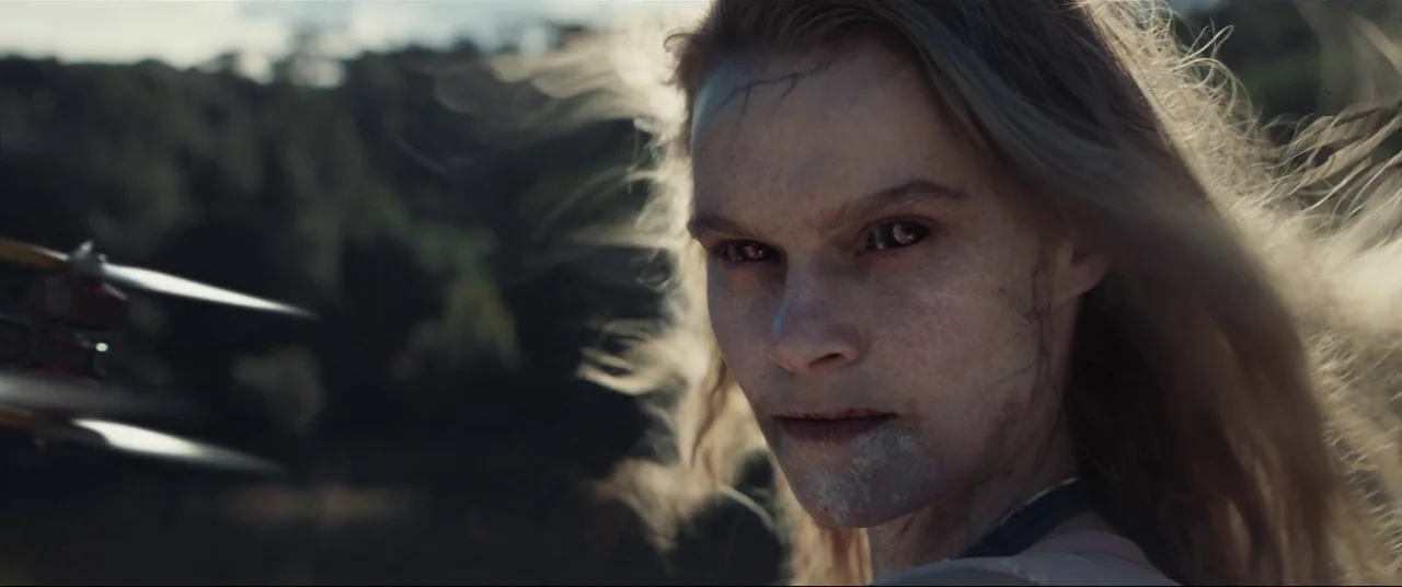 Evil Dead Rise (2023) movie still: closeup — A field with blurred trees in the background under daylight conditions. A young woman with black eyes…