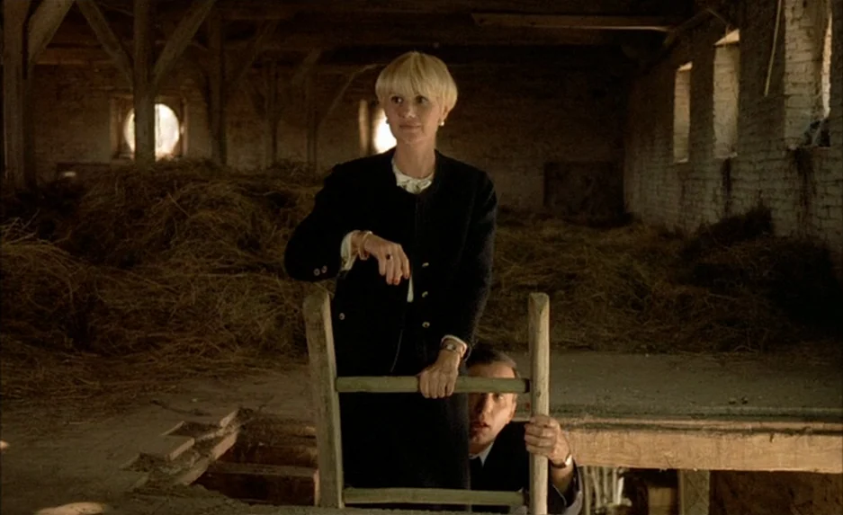 Milou en Mai (1990) movie still: medium — An interior barn loft with stacks of hay and exposed wooden ceiling beams. A woman stands on a ladder…