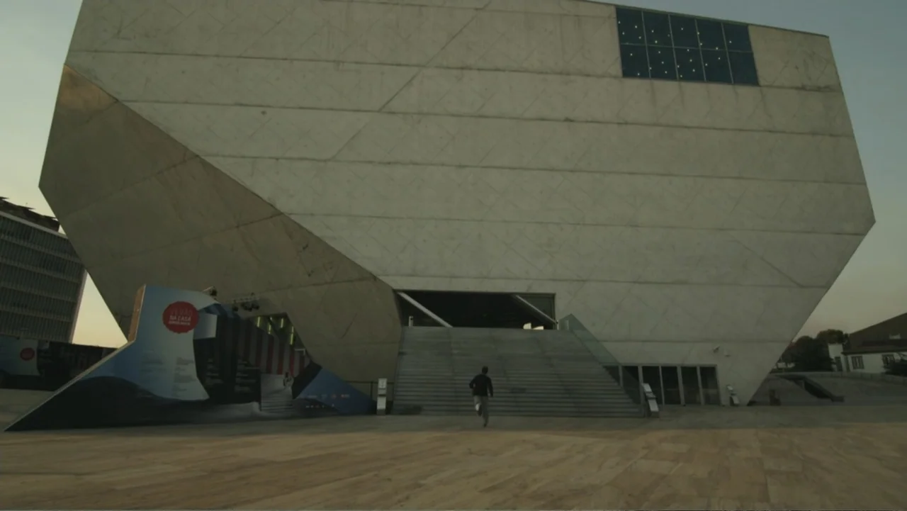 REM (2019) movie still: wide — A large, modern building with geometric concrete walls features a grand staircase at dusk. A person in dark clothing…