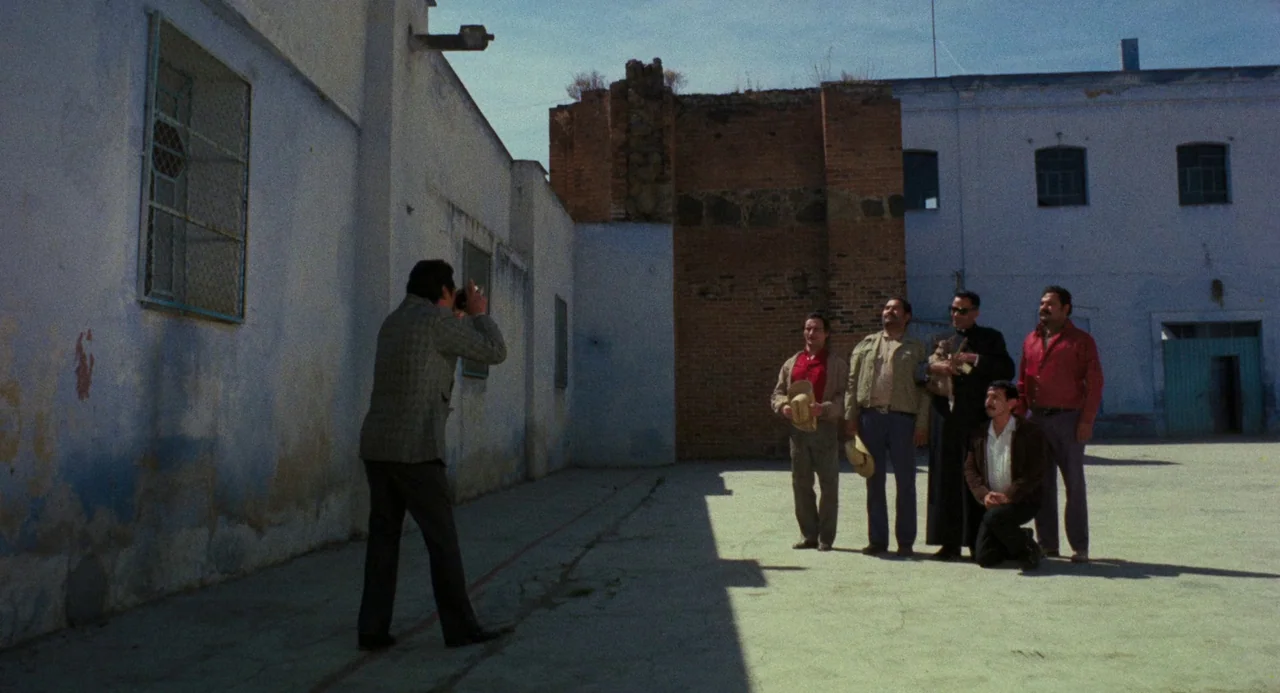 Canoa: A Shameful Memory (1976) movie still: wide — A sun-drenched courtyard with pale blue walls and an exposed brick facade behind. Six men are…