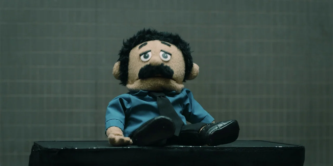 Música (2024) movie still: medium — A puppet with a mustache sits on a black platform against a grey grid background. The puppet wears a blue…