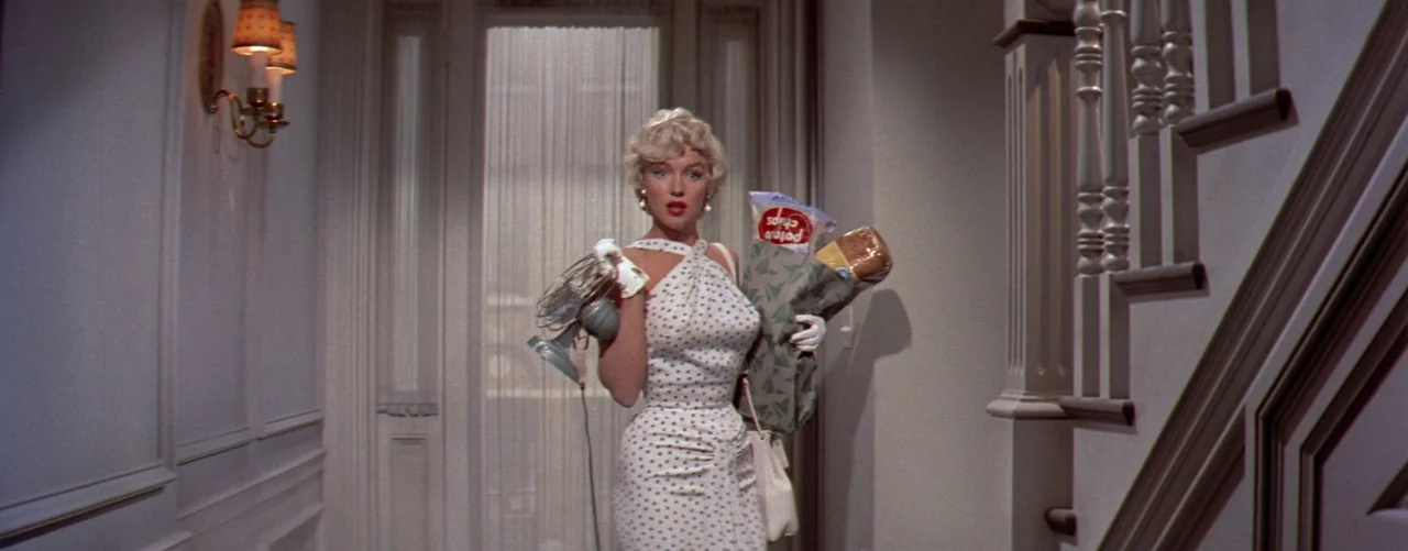 The Seven Year Itch (1955) movie still: fullbody — An interior hallway features a central door with a sheer curtain and a staircase. A woman stands…