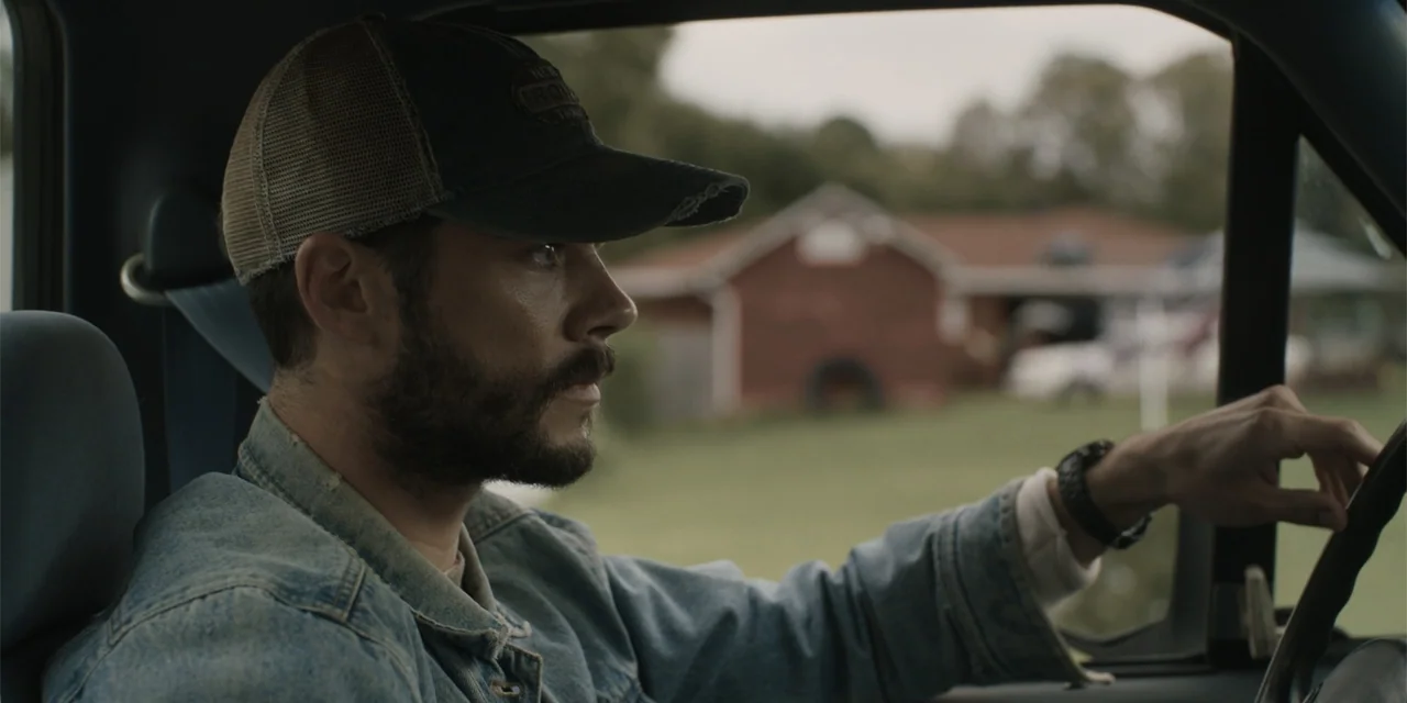 Caddo Lake (2024) movie still: medium — A man drives a truck through a rural landscape with a farm building visible outside. The man sits in the…
