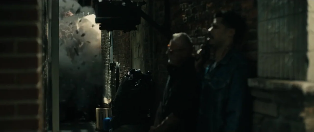 Guns Up (2025) movie still: medium — A narrow dark passage features exposed brick walls and a large cloud of debris. Two men stand in the foreground…