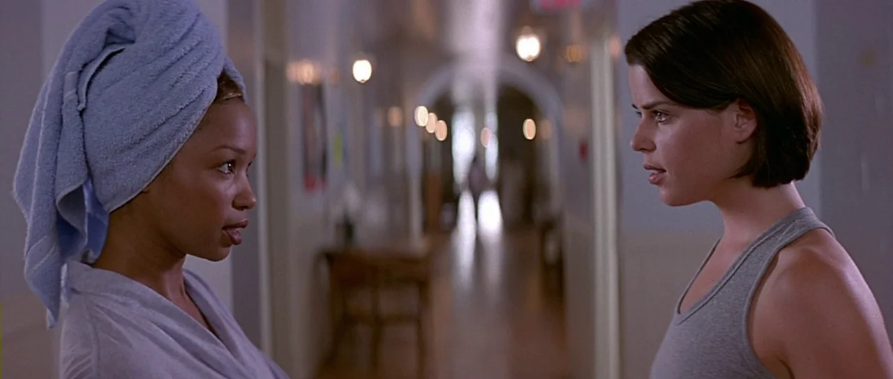 Scream 2 (1997) movie still: medium — A long indoor corridor with pale walls and wooden flooring leads to a window. Two young women stand facing…