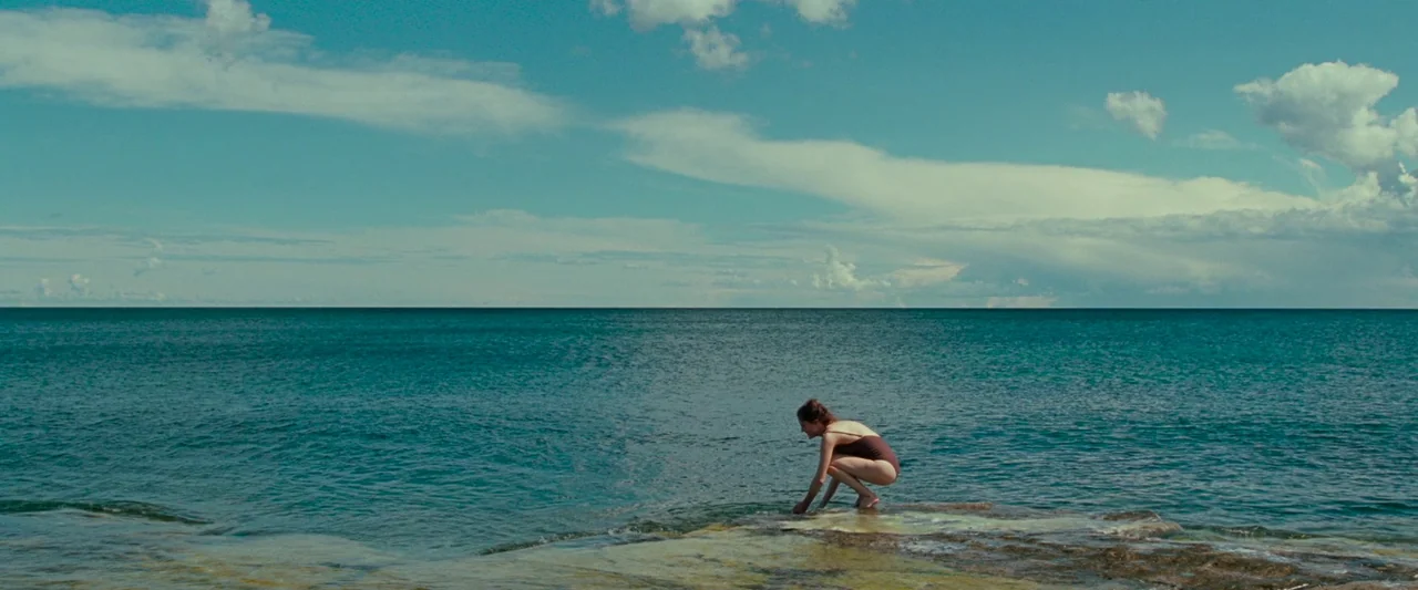 Bergman Island (2021) movie still: wide — A flat rocky shoreline extends into a vast calm blue sea under a sky. A woman in a swimsuit crouches on…