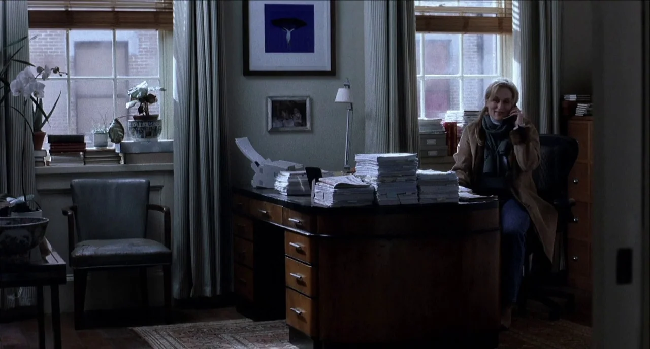 The Hours (2002) movie still: wide — A residential room featuring a large wooden desk filled with stacks of paper documents. A woman sits in a chair…