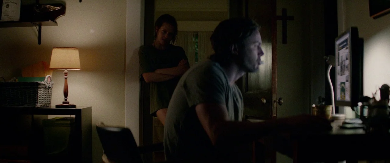 Triple 9 (2016) movie still: medium — A dark, cluttered interior room with a wooden door featuring a wall-mounted cross. A man sits at a desk using…
