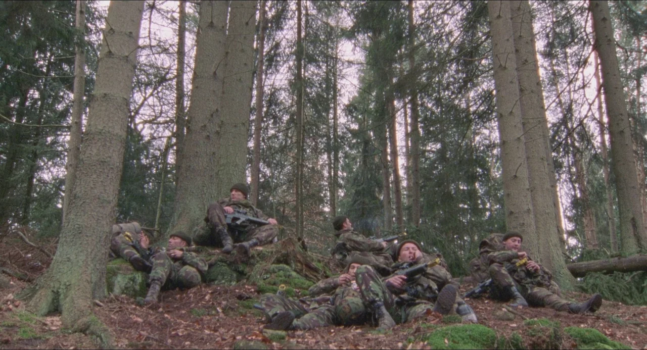 Dog Soldiers (2002) movie still: wide — A group of soldiers rests among large pine trees on a mossy, leaf-covered slope. The men sit and lounge on…