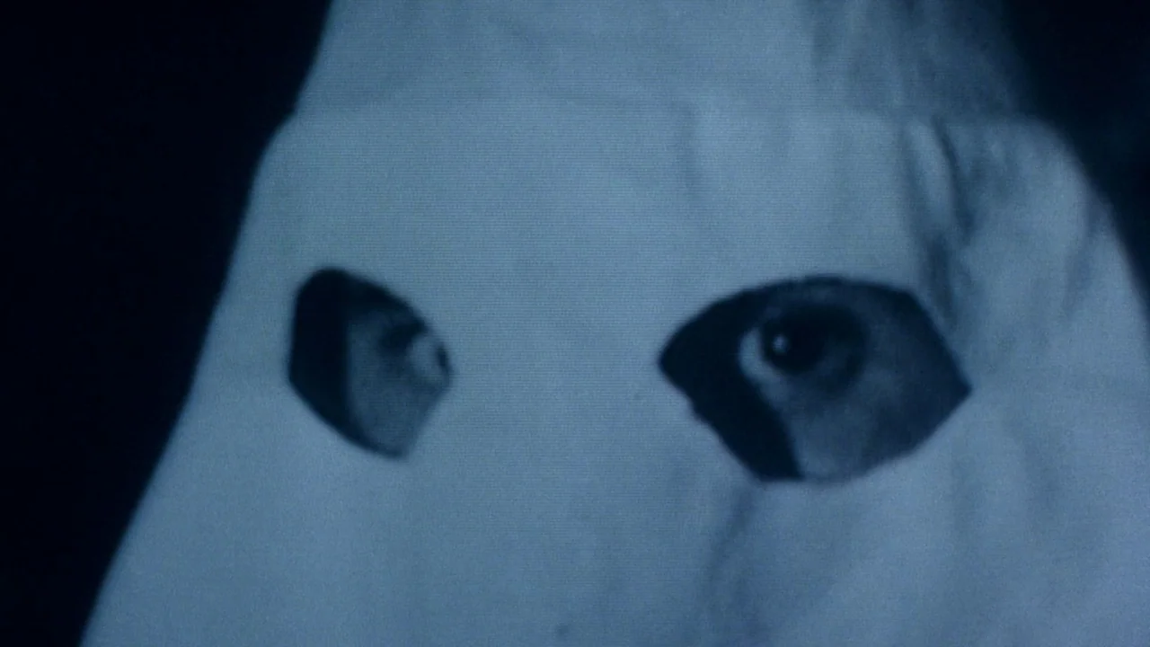 Malcolm X (1992) movie still: closeup — A close-up view shows a person wearing a white hood with cut-out eye holes. The person stares directly…