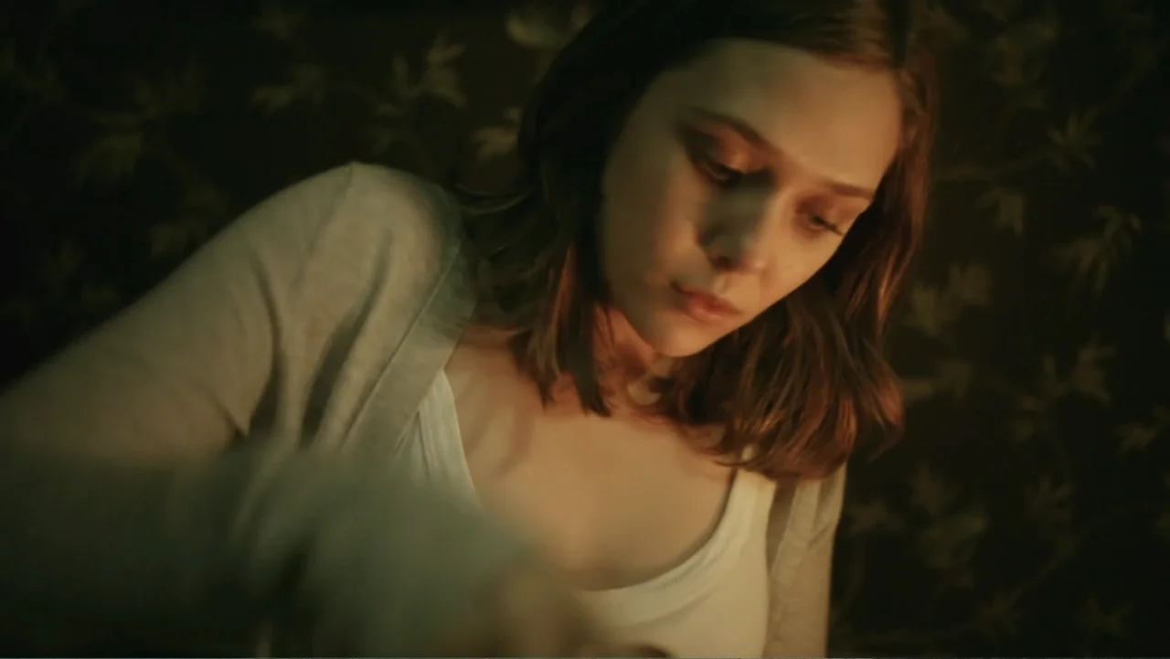 Silent House (2011) movie still: medium — An interior bedroom space features dark wallpaper with a repeating leaf pattern. A young woman looks…