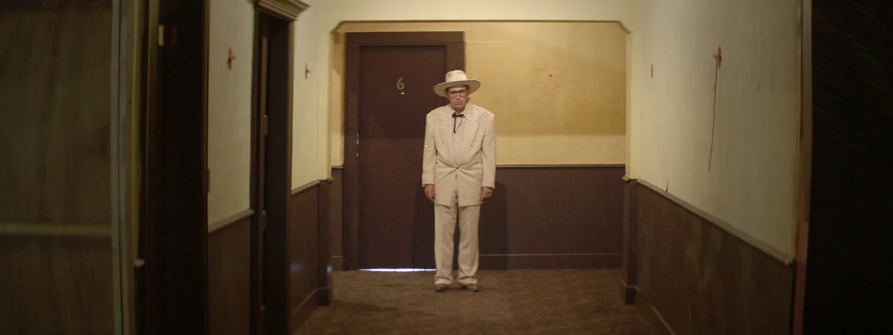 Entertainment (2015) movie still: fullbody — A narrow motel hallway features cream walls above dark brown wood paneling. An elderly man in a cream…