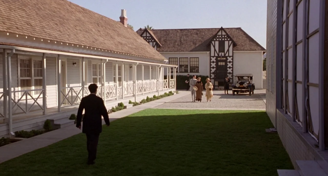 Chaplin (1992) movie still: wide — A well-manicured lawn separates two white wooden buildings with architectural details from the early 1900s. A man…