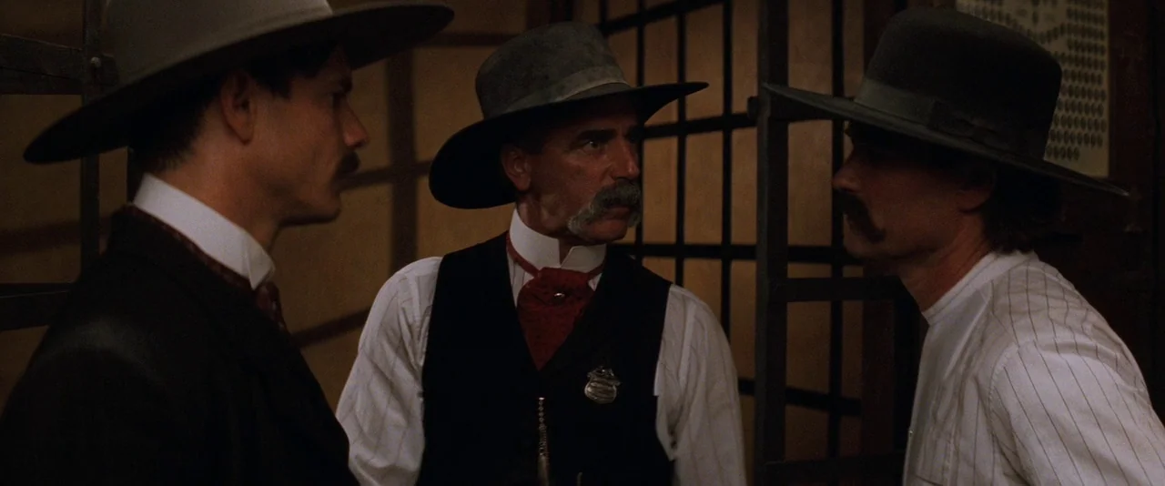 Tombstone (1993) movie still: medium — An interior space features wood-slat walls with dark iron bars dividing the area. Three men wearing period…