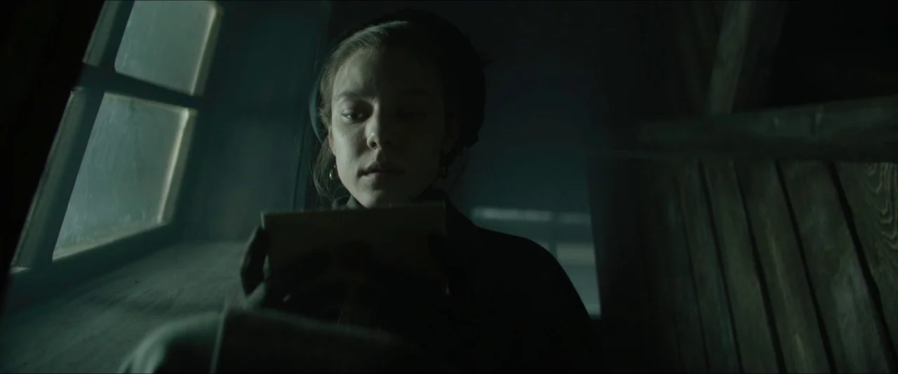 Tchaikovsky’s Wife (2022) movie still: closeup — A dimly lit attic with wooden walls is illuminated by daylight from a window. A young woman…