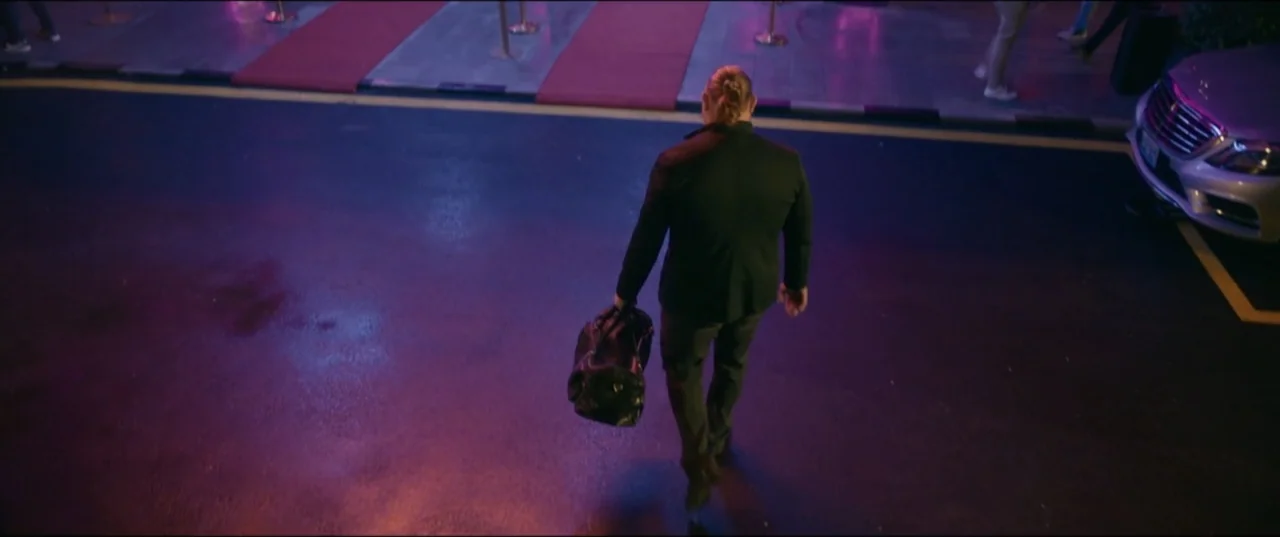 Risqué (2025) movie still: fullbody — A man walks across a dark asphalt lot at night near a red carpet entrance. He wears a black suit and carries a…