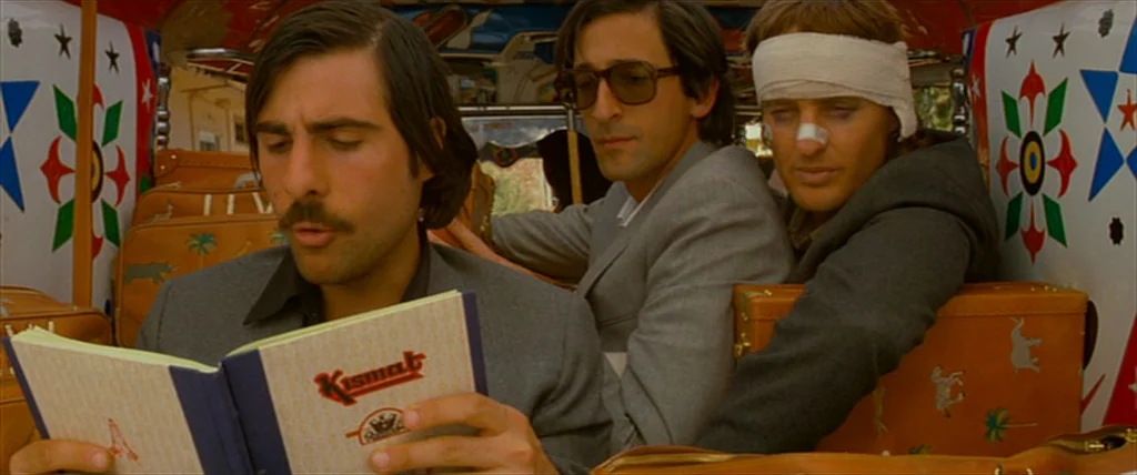 The Darjeeling Limited (2007) movie still: medium — Three men sit inside a brightly decorated auto rickshaw with patterned surfaces and luggage. The…