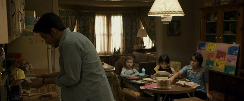 Zodiac (2007) movie still: medium — A man in a blue shirt works at a counter in a home interior. Three children sit at a small table covered with…