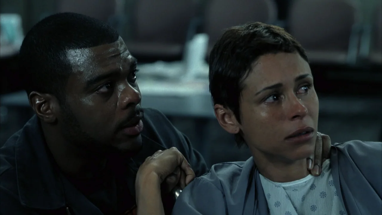 John Q (2002) movie still: medium — A dimly lit hospital waiting area features rows of out-of-focus chairs in the background. A man sits beside a…