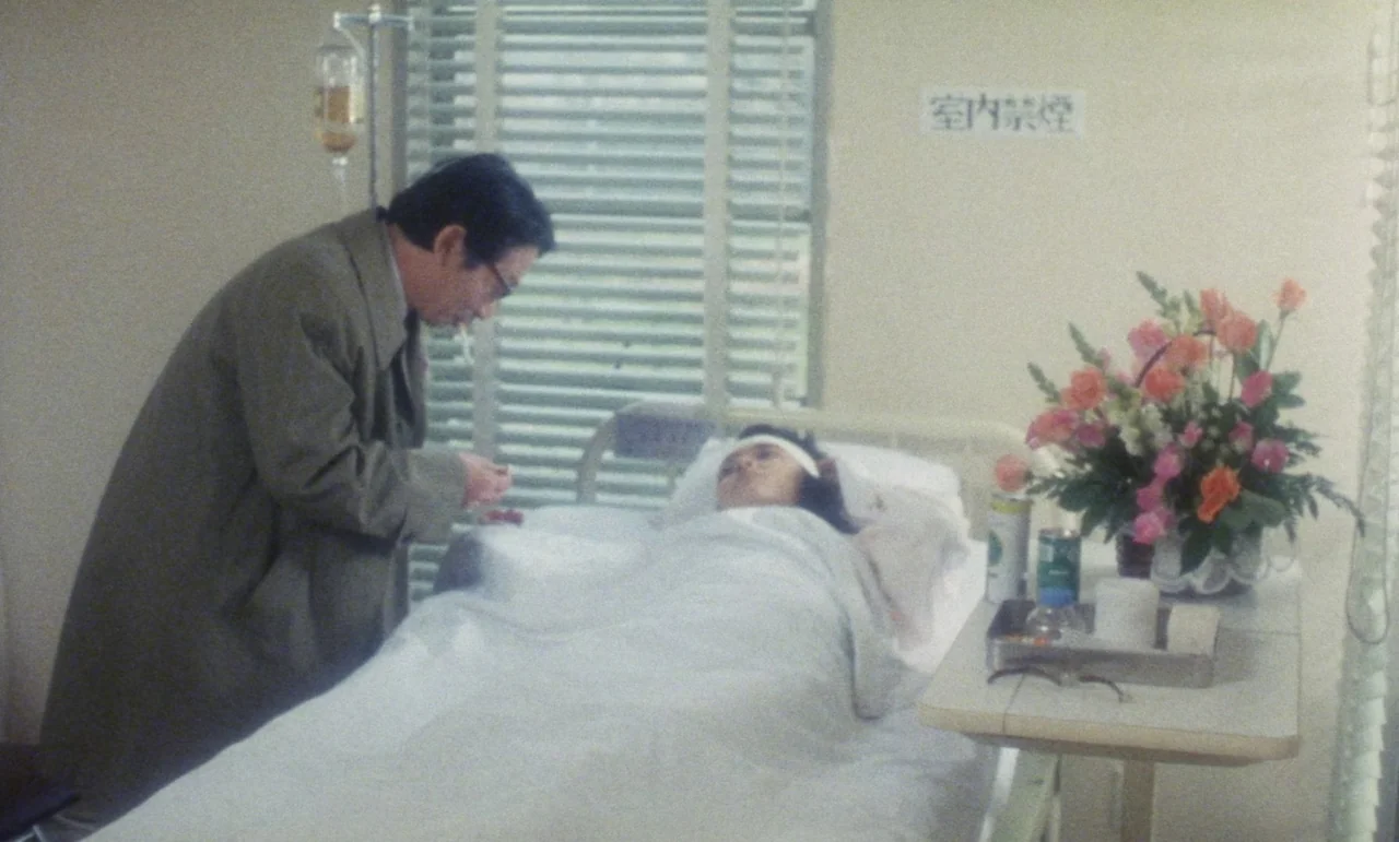 Evil Dead Trap (1988) movie still: medium — A hospital room features white walls, metal window blinds, and a patient bed. A woman with a head…