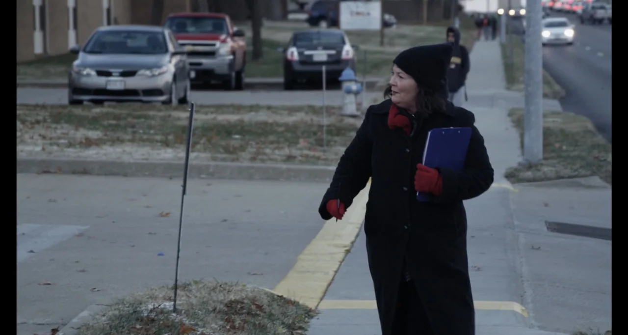 Abortion: Stories Women Tell (2016) movie still: fullbody — A suburban sidewalk runs alongside a paved road with parked cars and distant traffic. A…
