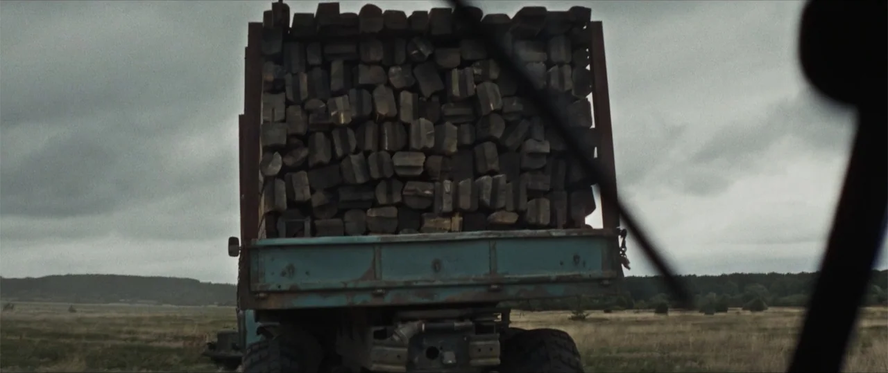 Sisu: Road to Revenge (2025) movie still: medium — A rural expanse features a weathered flatbed truck carrying a heavy load of logs. The perspective…
