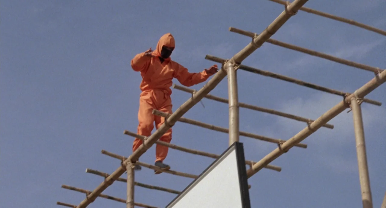 American Ninja (1985) movie still: fullbody — A person stands on a high bamboo structure against a clear sky. The subject balances on the wooden…