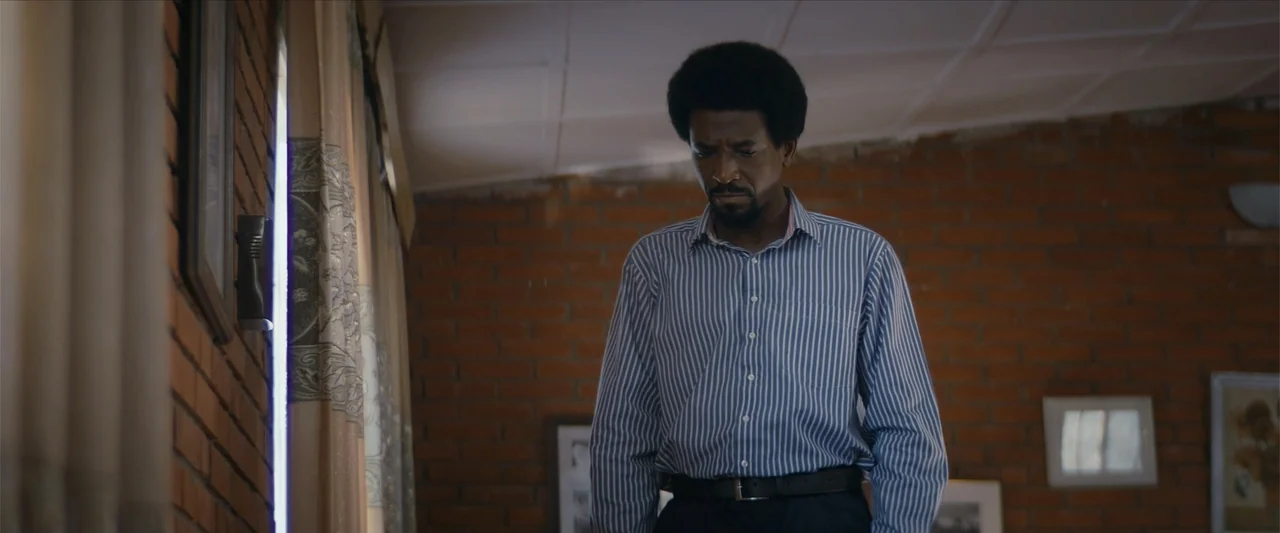 Ms. Kanyin (2025) movie still: medium — An interior space features exposed brick walls and a window with light beige drapes. A man in a striped…