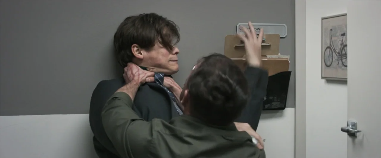 Office Uprising (2018) movie still: medium — An office hallway with a grey wall and a framed bicycle art piece. One man pins another man against the…