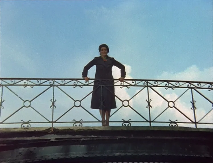 Claire’s Knee (1970) movie still: fullbody — A woman stands on an arched metal bridge with an intricate decorative railing. The blue sky fills the…