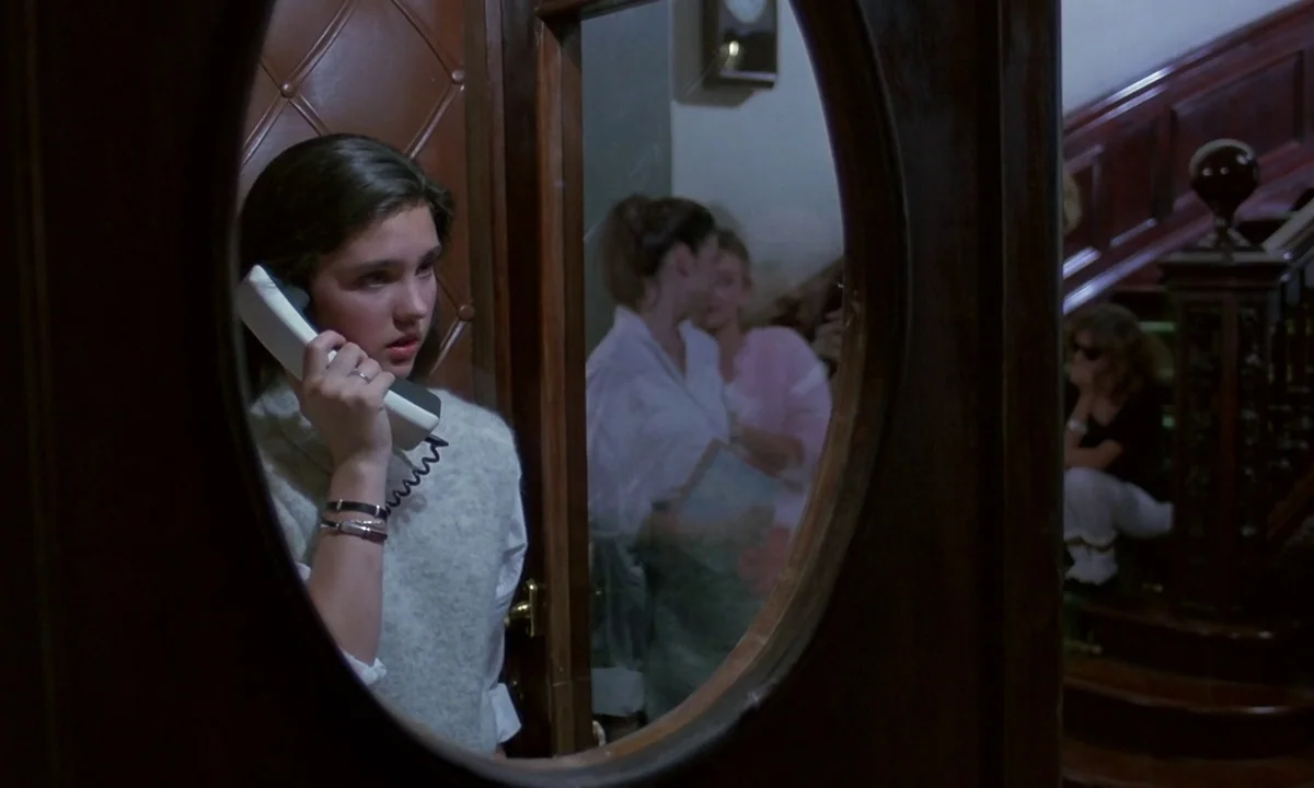 Phenomena (1985) movie still: medium — A woman stands in a wooden-framed doorway while holding a white telephone receiver. In the background, two…