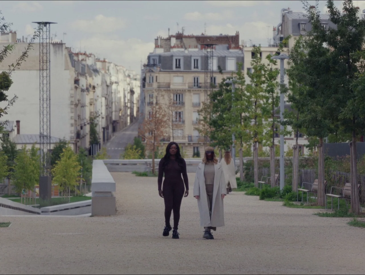 Transformers: Rise of the Beasts (2023) movie still: fullbody — An urban pedestrian walkway surrounded by young trees and residential Parisian-style…