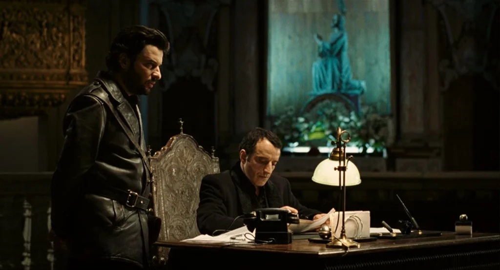 This Night (2008) movie still: medium — A dark wooden desk sits before a large, illuminated religious painting in an ornate cathedral. One man…