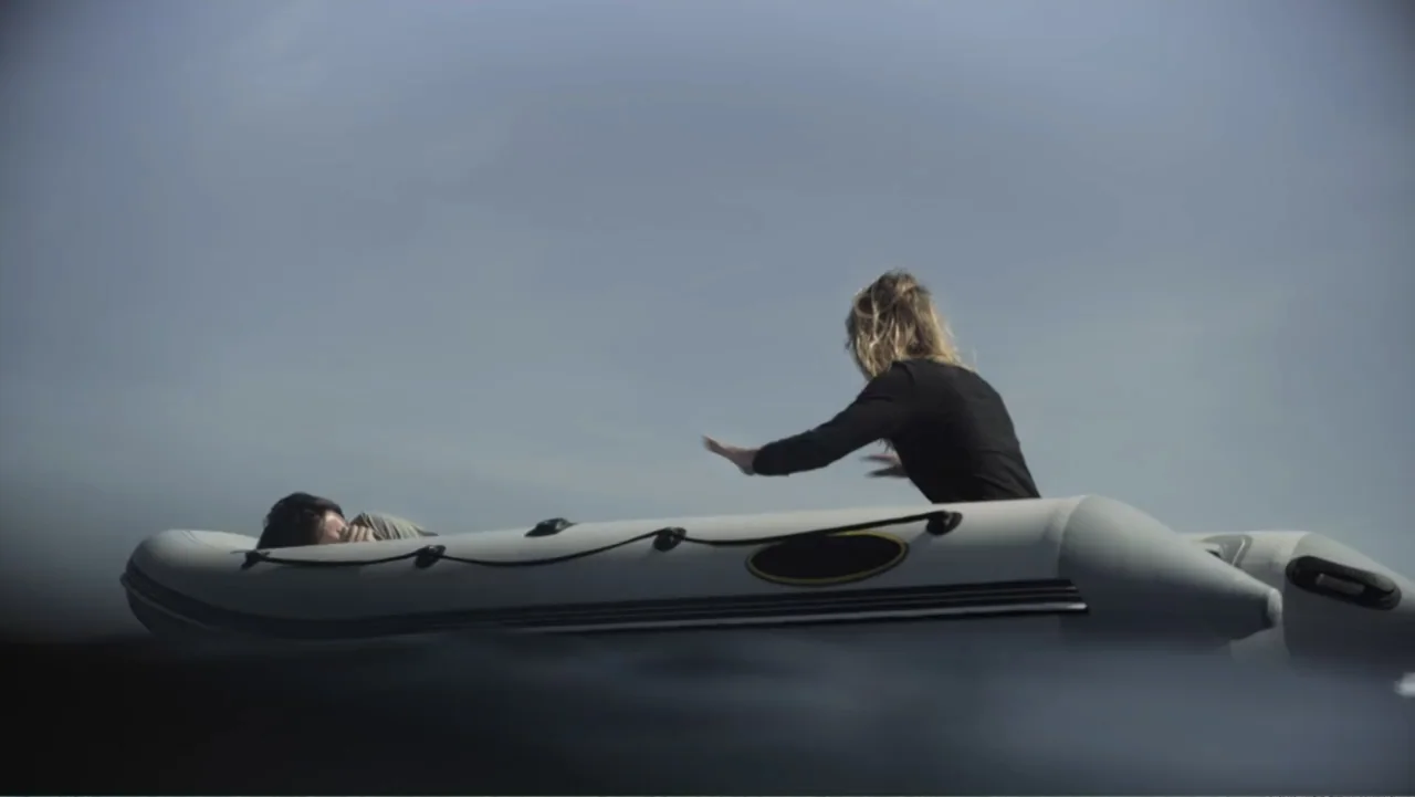 Capsized: Blood in the Water (2019) movie still: wide — An inflatable rubber raft floats on open water under a clear blue sky. A woman in a black…
