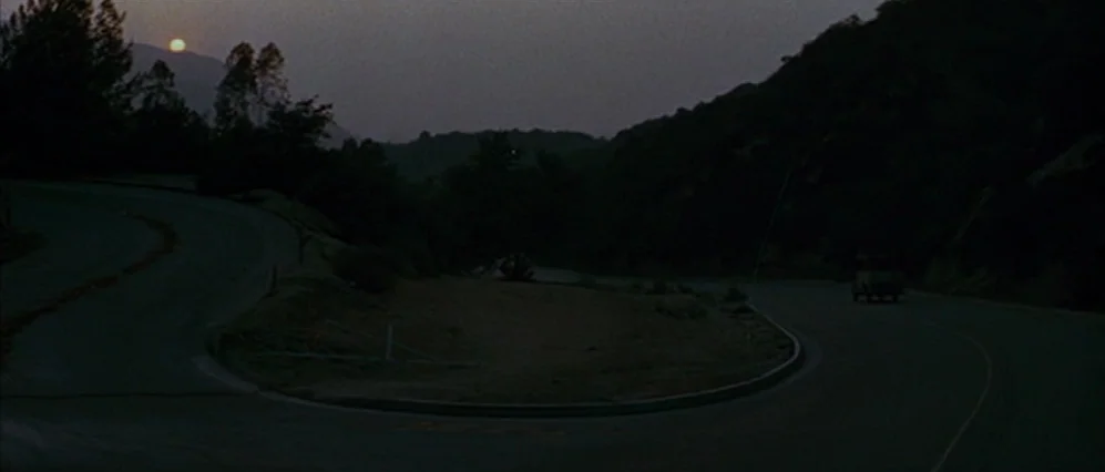 The Fortune (1975) movie still: wide — A winding asphalt road curves through a dark, hilly landscape at late sunset. A single small vehicle travels…