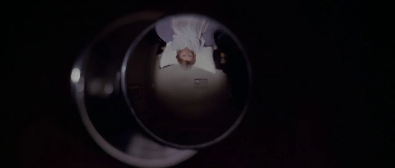 The New York Ripper (1982) movie still: closeup — A view through a circular door peephole showing a person in a darkened room. A woman lies in a bed…